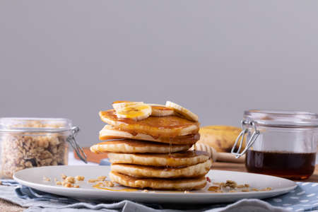 Pancakes with banana slices and syrup served in plate by oats and honey jar against gray background. unaltered, breakfast, copy space, sweet food, fruit and healthy food concept.の写真素材