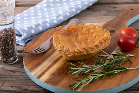 Stuffed pie with rosemary and tomatoes on serving board by black pepper in container at table....