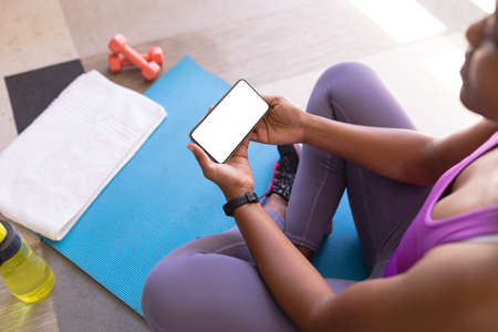 African american mid adult woman using smart phone while sitting on exercise mat at home, copy space. unaltered, wireless technology, home workout, fitness and active lifestyle concept.の写真素材