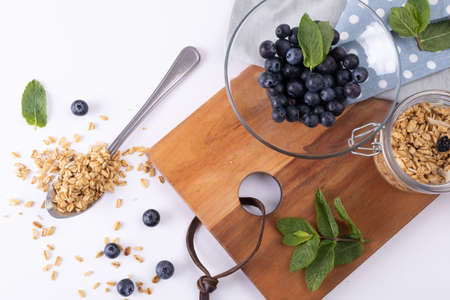 Close-up of blue berries in bowl with granola and herbs on serving board over white background....