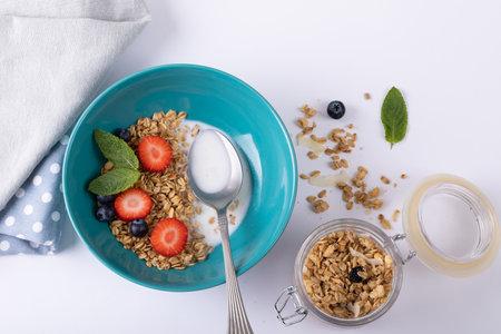 Directly above shot of granola with berry fruits, herbs and milk in bowl with spoon on table....