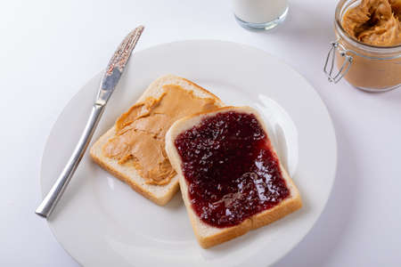Close-up of peanut butter and preserves on bread slices in plate with table knife, jar and milk....