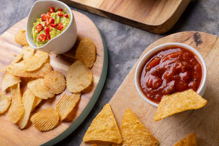 High angle view of potato chips and nacho chips with various salsa on serving board at table....