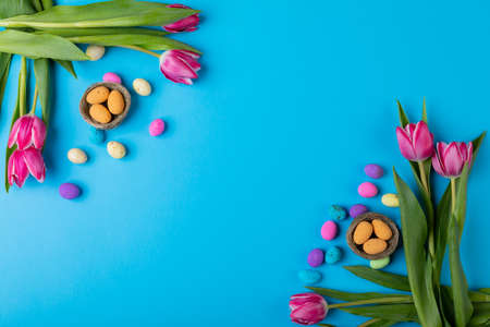Overhead view of easter candies in small nests with pink tulips on blue background with copy space....