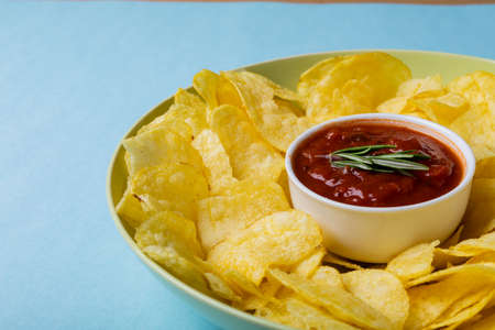Close-up of red sauce in bowl amidst potato chips served in plate on blue background. unaltered, unhealthy food, snack, dipping sauce, crunchy, salty snack and savory food.の写真素材