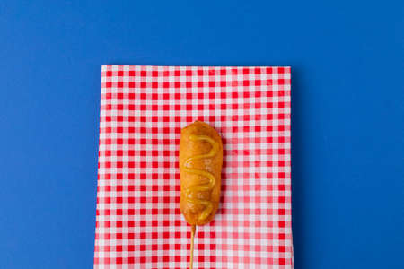 Directly above shot of corn dog with mustard sauce on checked pattern napkin over blue background....