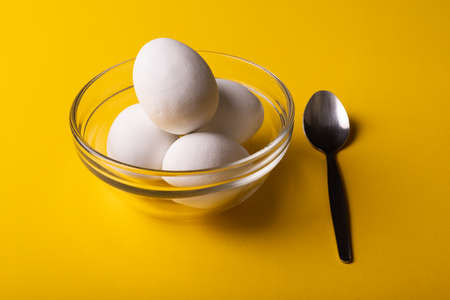 High angle close-up of fresh white eggs in glass bowl by spoon on yellow background. unaltered,...