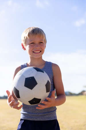 Smiling caucasian elementary schoolboy with soccer ball looking away while standing against sky. unaltered, childhood, education, sports training and sports activity concept.の写真素材