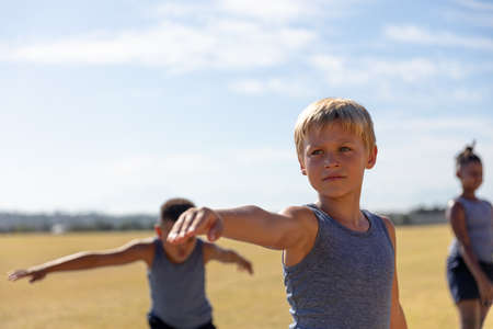 Caucasian elementary schoolboy with arm outstretched exercising against sky during sunny day....