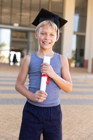 Portrait of happy caucasian elementary schoolboy with mortarboard and degree standing against school. unaltered, imitation, education, school and graduation concept.の写真素材