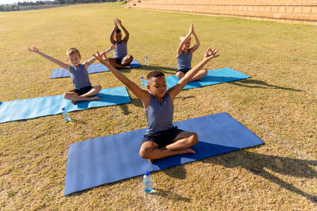 Multiracial elementary schoolboys with arms raised exercising while sitting on yoga mat in ground....