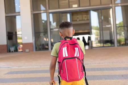 Rear view of biracial elementary schoolboy with backpack standing against school building. unaltered, childhood, education and back to school concept.の写真素材