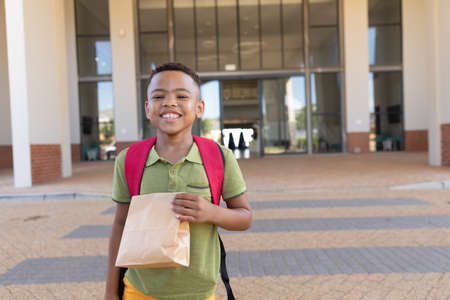 Portrait of smiling biracial elementary schoolboy with packed lunch standing in school campus. unaltered, childhood, education and back to school concept.の写真素材