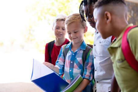 Caucasian elementary schoolboy showing books to multiracial male classmates at school. unaltered,...
