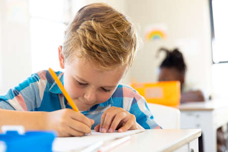 Close-up of cute caucasian elementary schoolboy writing on book while sitting at desk in school. unaltered, childhood, education, learning, writing and back to school concept.の写真素材