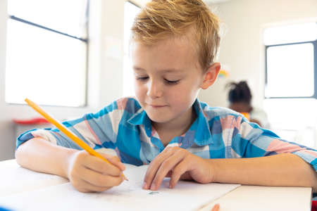 Close-up of caucasian elementary schoolboy writing on book while sitting at desk in school....