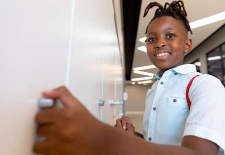 Portrait of smiling african american elementary schoolboy standing by locker in school. unaltered,...