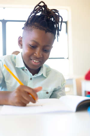 Smiling african american elementary schoolboy writing on book while sitting at desk in school. unaltered, childhood, education, learning, writing and back to school concept.の写真素材