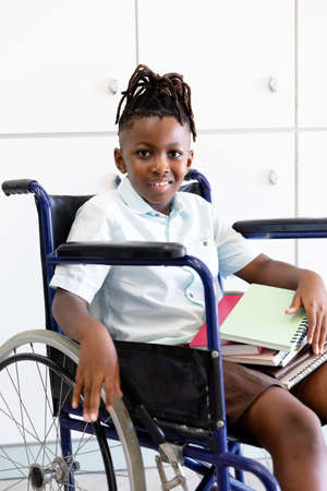 Portrait of african american elementary schoolboy with books sitting on wheelchair in corridor. unaltered, childhood, disability, physical disability, education and back to school concept.の写真素材