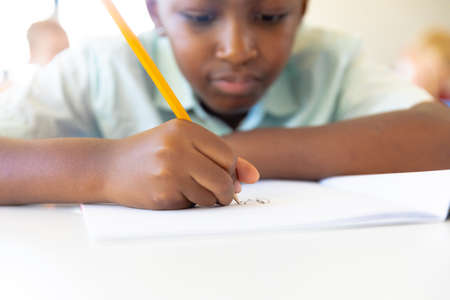 Close-up of african american elementary schoolboy writing on book at desk in classroom. unaltered,...