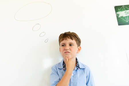 Thoughtful caucasian elementary schoolboy with hand on chin standing by speech bubble on wall. unaltered, childhood, education and back to school concept.の写真素材