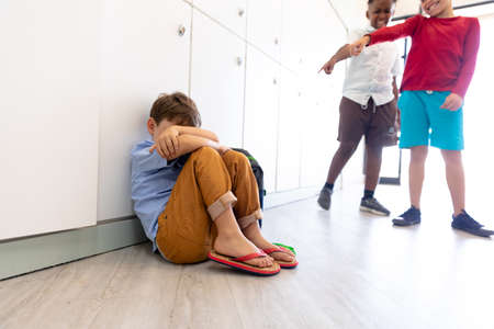 Multiracial elementary schoolboys laughing while gesturing over sad male classmate sitting on floor. unaltered, childhood, bullying, problems, sadness, cruel, education and back to school concept.の写真素材
