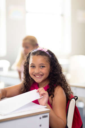 Portrait of smiling cute biracial elementary schoolgirl with book sitting at desk in classroom....