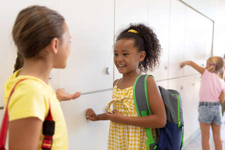 Happy multiracial elementary schoolgirls talking while standing by lockers in school corridor....