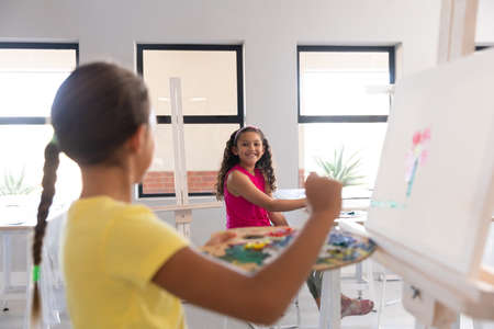 Smiling multiracial elementary schoolgirls painting on easel during drawing class in school. unaltered, childhood, education, art, painting, activity and back to school concept.の写真素材