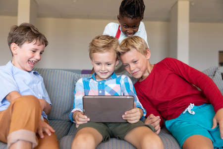 Smiling multiracial elementary schoolboys looking at digital tablet while sitting on couch in...