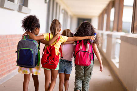 Rear view of multiracial elementary schoolgirls with backpacks and arm around walking in corridor. unaltered, childhood, together, education and back to school concept.の写真素材