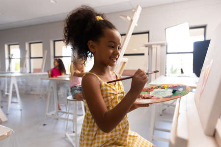 Smiling biracial elementary schoolgirl with palette and paintbrush painting on easel in school. unaltered, childhood, education, curly hair, art, painting, activity and back to school concept.の写真素材