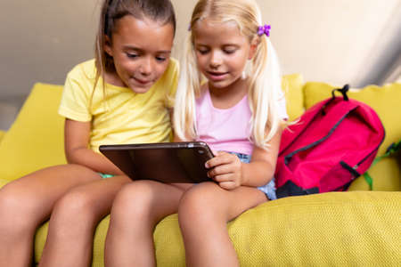 Smiling caucasian elementary schoolgirls looking at digital tablet while sitting on couch in school. unaltered, childhood, wireless technology, together, education and back to school concept.の写真素材