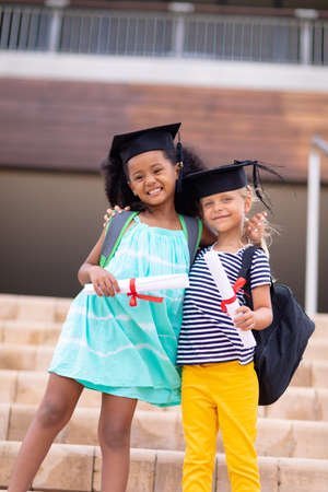 Portrait of smiling multiracial elementary schoolgirls with mortarboard and degree standing on steps. unaltered, imitation, happiness, together, education, graduation and school concept.の写真素材