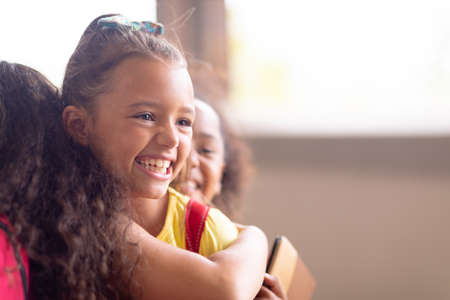 Close-up of multiracial cheerful elementary schoolgirls embracing while standing in school. unaltered, childhood, education and back to school concept.の写真素材