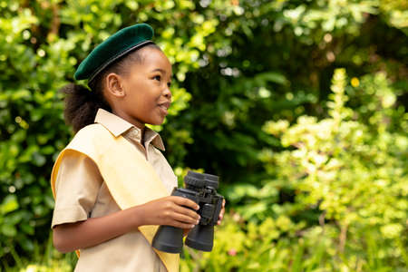 Smiling african american scout girl in uniform holding binoculars in forest. unaltered, girl scout, childhood, exploration, discovery, adventure and scouting.の写真素材