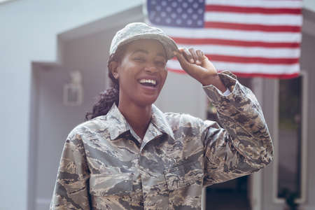 Happy female mid adult african american soldier in uniform standing outside house. unaltered, pride, military, flag of america, homecoming, armed forces and patriotism concept.の写真素材