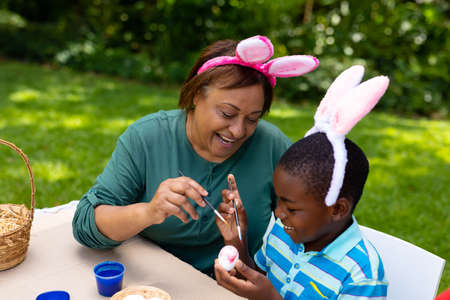 Smiling african american boy and grandmother in bunny ears painting egg at backyard on easter day....