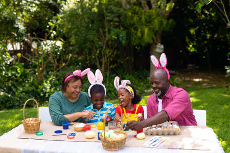 Cheerful african american siblings and grandparents in bunny ears painting eggs on easter day. unaltered, lifestyle, easter day, art, celebration, family, cultures and holiday concept.の写真素材