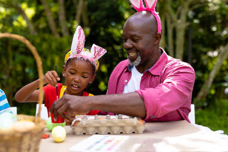 African american girl and grandfather wearing bunny ears while painting eggs on easter day....