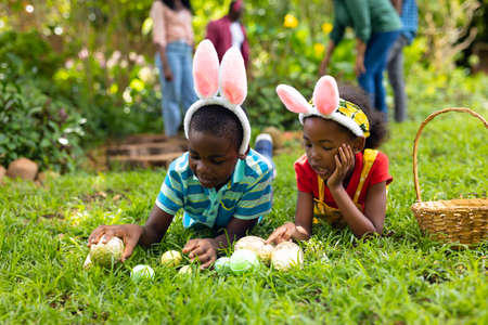 African american siblings in bunny ears arranging easter eggs on grass while family in background. unaltered, lifestyle, easter day, celebration, childhood, cultures and holiday concept.の写真素材