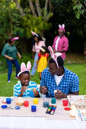 Happy african american boy and father painting easter eggs while family in background. unaltered,...