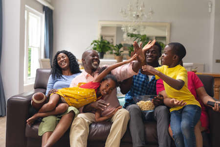Happy african american multi-generational family high-fiving while watching rugby match on tv. unaltered, lifestyle, sport, fans, family, watching tv, rugby match, cheering and leisure concept.の写真素材