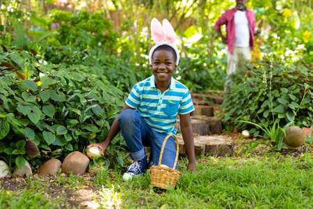 Portrait of smiling cute african american boy wearing bunny ears hiding easter egg in backyard....