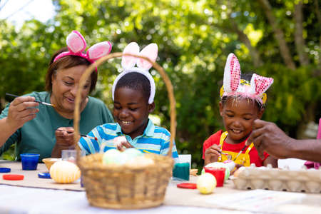 Smiling african american siblings and grandmother wearing bunny ears while painting eggs on easter....