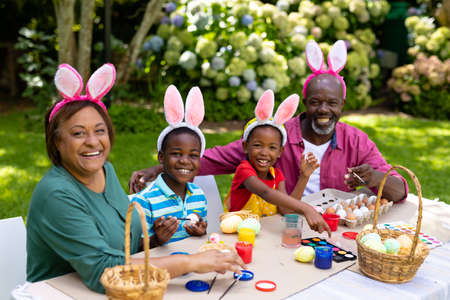 Portrait of happy african american siblings and grandparents in bunny ears painting easter eggs....