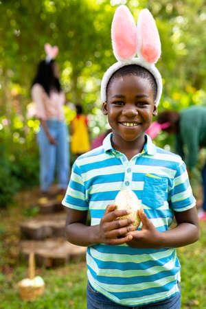 Portrait of smiling african american boy in bunny ears holding easter egg while family in backyard....