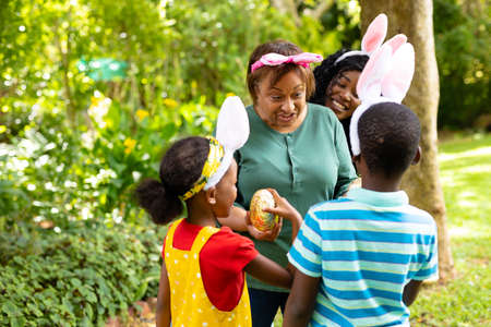 African american siblings in bunny ears giving easter egg to grandmother in backyard. unaltered,...