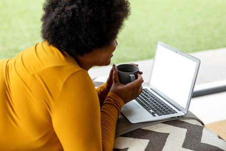 Side view of african american mid adult woman holding coffee cup while using laptop at home....