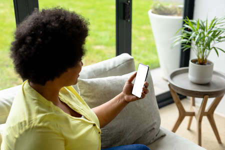 African american mid adult woman using smart phone with blank screen while sitting on sofa at home....